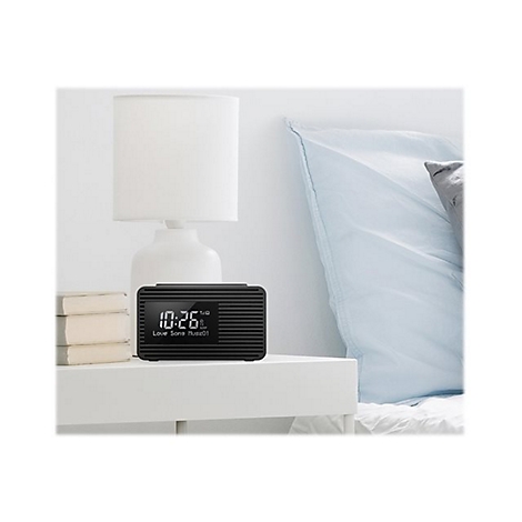 Schwarzer Radiowecker mit Digitalanzeige (10:26) auf einem Nachttisch neben einer Lampe und Büchern, vor einem Bett.