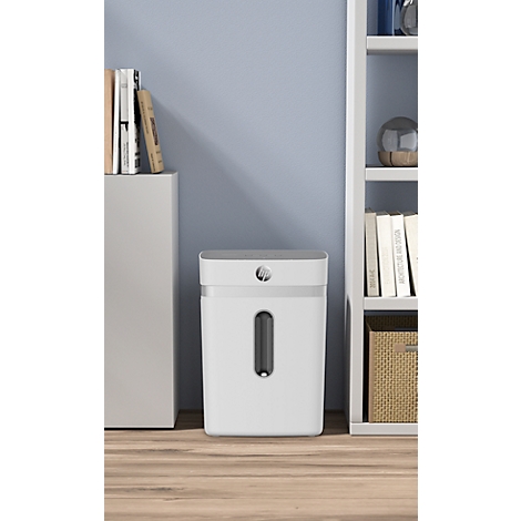 White paper shredder with viewing window, standing on light wooden floor in front of light blue wall. Books to the left, shelf with books to the right.