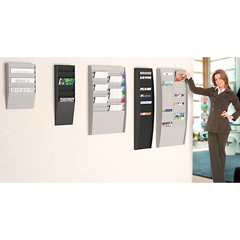 A woman stands beside several wall-mounted brochure holders. Various sizes and colors, filled with brochures and papers.