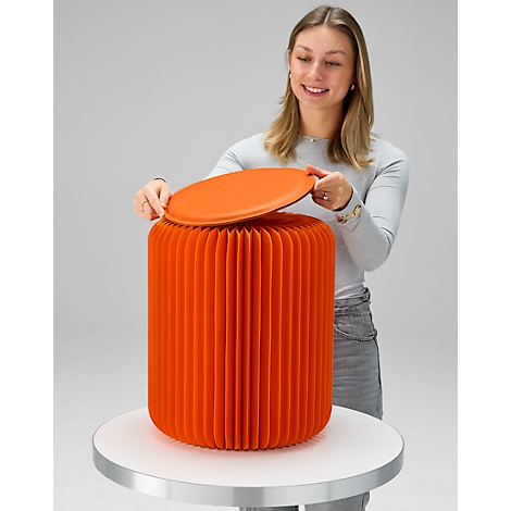 Frau öffnet orangenen, faltbaren Hocker, der auf einem Tisch steht.