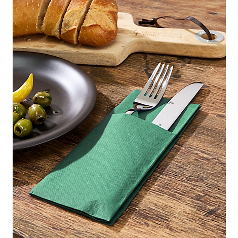 Besteck in grüner Serviette auf Holztisch. Links Teller mit Oliven, Brot und Schneidebrett im Hintergrund.
