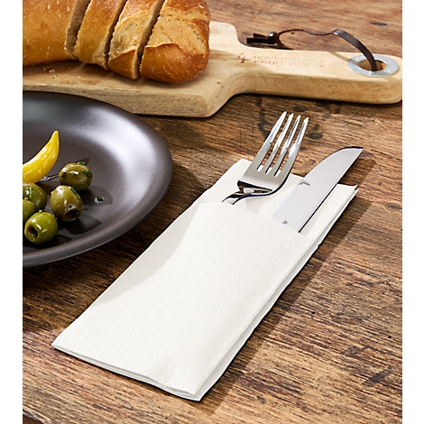 Besteck in Papiertasche auf Holztisch. Links Teller mit Oliven, Brot und Schneidebrett im Hintergrund.