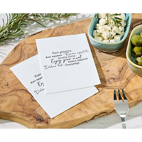Zwei Servietten mit Aufdrucken von 'Buen proveche, Bon appétit, Enjoy your meal' auf einem Holzbrett. Dazu Oliven, Käse und eine Gabel.