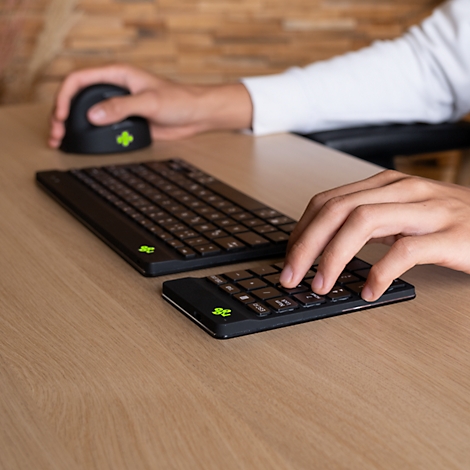 Une personne utilise un clavier, un pavé numérique et une souris sur une surface en bois. Tous les appareils sont noirs avec un logo vert.