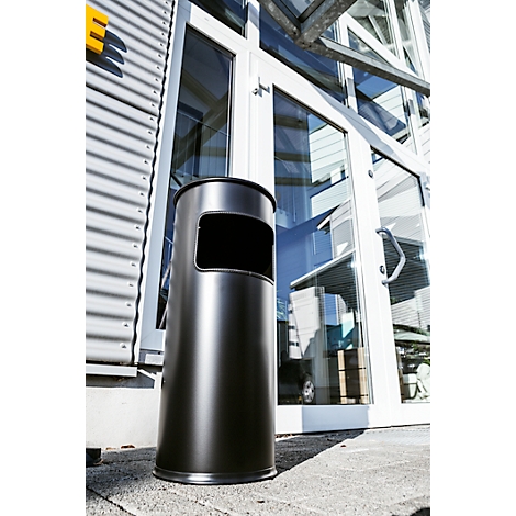 Black trash can in front of glass facade. The bin has a rectangular opening. Buildings and sky can be seen in the background.
