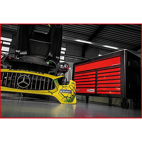 Gelbe Mercedes Stoßstange mit Michelin Aufkleber vor roter Werkzeugkiste. Schwarzer Wagenheber und offene Motorhaube.