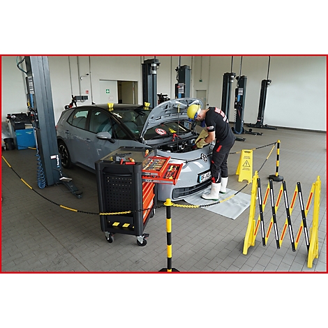 Ein Mechaniker repariert ein graues Auto in einer Werkstatt. Er trägt eine gelbe Schutzhaube und Arbeitskleidung.