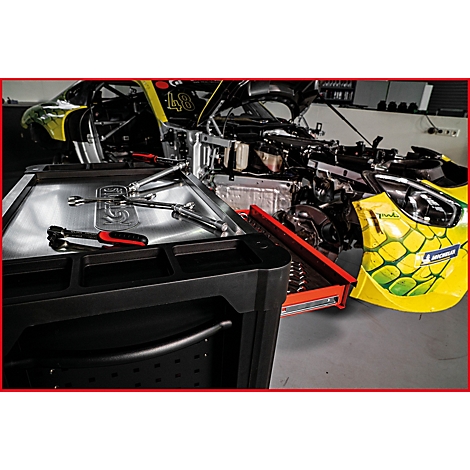 Ein Werkzeugwagen mit Werkzeugen vor einem teilweise demontierten Rennwagen. Im Hintergrund ist ein gelbes Michelin-Tuch zu sehen.