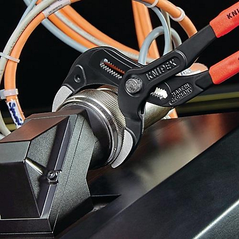 Une pince KNIPEX noire agrippe un objet cylindrique argenté. Câbles et pièces de machines en arrière-plan.