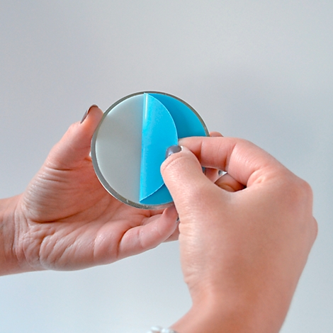 Des mains tiennent un disque rond et transparent, en train de décoller un film protecteur bleu. Le fond est blanc.