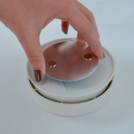 Une personne place une plaque métallique sur un objet rond blanc. Plaque avec deux aimants, main avec vernis à ongles foncé.
