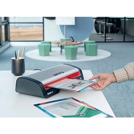 Une personne plastifie un certificat avec une plastifieuse grise et rouge. D'autres documents sont sur la table.