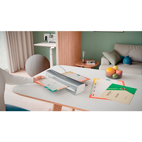Personne laminant du papier, avec d'autres feuilles et des fruits sur une table. Arrière-plan : canapé, bureau debout.