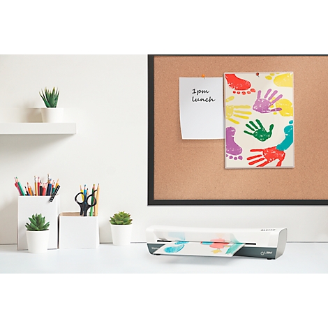 Une plastifieuse sur un bureau. Sur un tableau en liège, une feuille avec des empreintes de mains et de pieds colorées. À gauche, crayons, ciseaux et petites plantes.