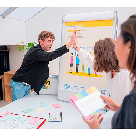 Un homme et une femme se font un high-five devant un tableau à feuilles mobiles. Une troisième personne tient des notes. Il y a des post-it sur la table.