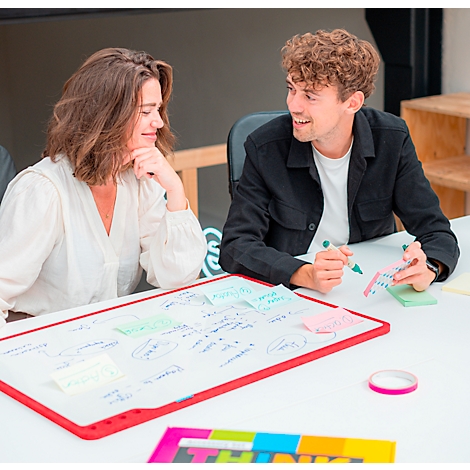 Deux personnes sont assises à une table avec un tableau blanc, en discussion. Il y a des notes et des marqueurs sur la table.