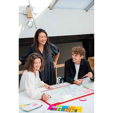 Trois personnes souriantes sont assises à une table en train de travailler sur un projet. Des notes, des post-it colorés et des stylos sont visibles.