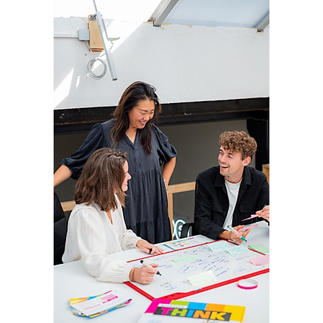 Trois personnes sont assises autour d'une table et travaillent ensemble sur une présentation. Une femme se tient à côté, souriante.