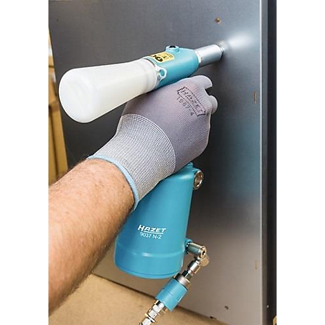Een persoon houdt een blauwe pneumatische tool tegen een metalen plaat. De handschoen draagt het HAZET-logo. De achtergrond is wazig.