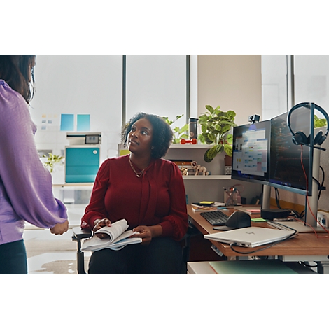 A woman in a red blouse is seated, looking up at a person standing in front of her, holding a book. A desk with computers is visible.