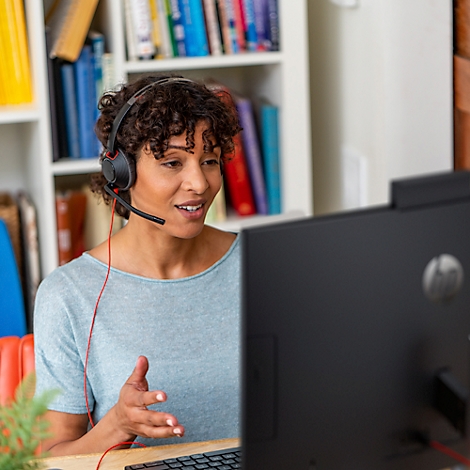 Woman with headset sits in front of a computer, gesturing. Bookshelf in the background.