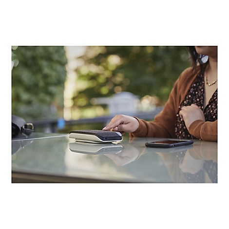 Eine Frau legt ihr Smartphone neben einen Lautsprecher auf einen Tisch. Im Hintergrund ist ein Baum unscharf.