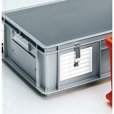 Caja de plástico gris con tapa, etiqueta de dirección y cierre metálico. Partes de una caja roja se pueden ver en el fondo.