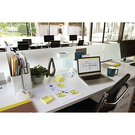 Espace de travail de bureau avec ordinateur portable, notes et plantes. En arrière-plan, d'autres espaces de travail et une personne.