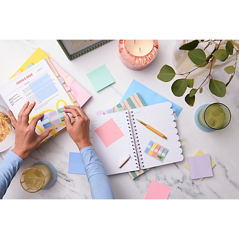 Vue de dessus d'une main prenant des notes dans un cahier. Des stylos, des post-it et des boissons sont éparpillés autour.