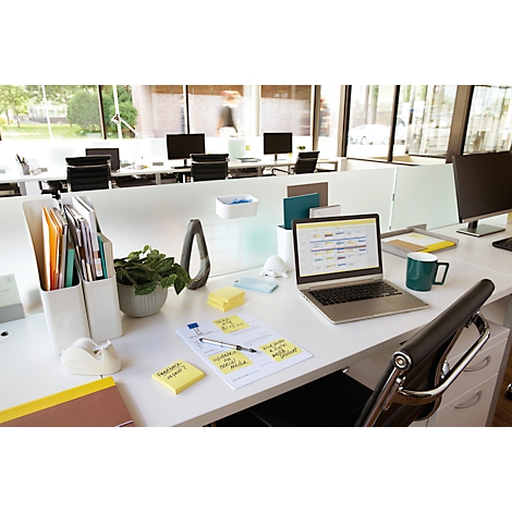 Büroarbeitsplatz mit Laptop, Notizen, Stiften, grüner Tasse und Pflanze. Im Hintergrund weitere Arbeitsplätze.
