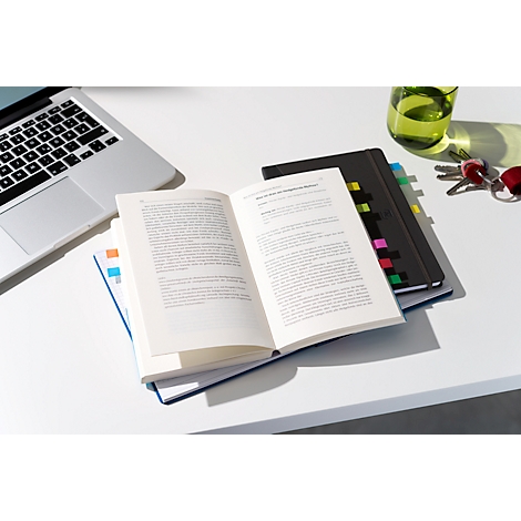 Livre ouvert avec des notes adhésives colorées, un ordinateur portable, un verre et des clés sur une table blanche.