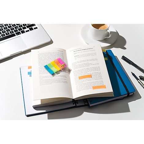 Livre ouvert avec des post-it colorés, un ordinateur portable, une tasse de café et des stylos sur un bureau blanc.