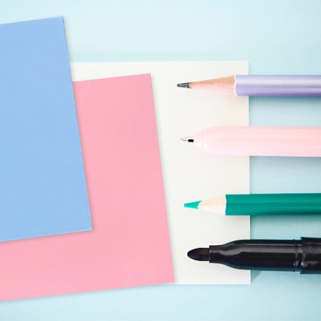 Gekleurde pennen en papier gedrapeerd op een lichtblauwe achtergrond. Paars potlood, roze pen, groene potlood, zwarte stift.