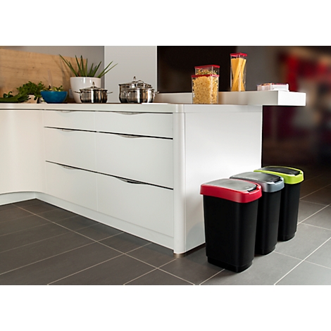 Trois poubelles noires avec des couvercles colorés sont dans une cuisine. L'arrière-plan montre une cuisine blanche avec des casseroles argentées.