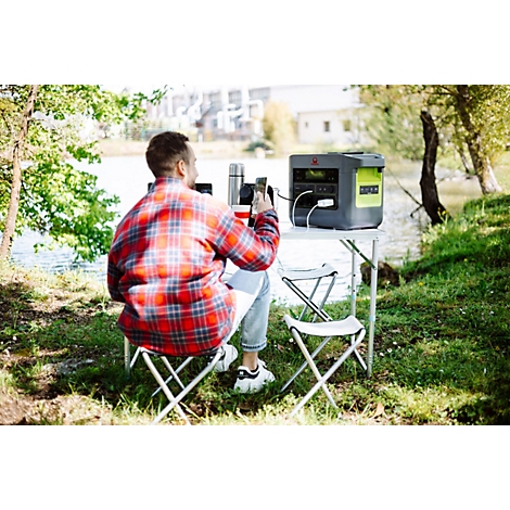 Ein Mann sitzt mit einem Smartphone an einem Campingtisch am See und nutzt eine tragbare Powerstation.