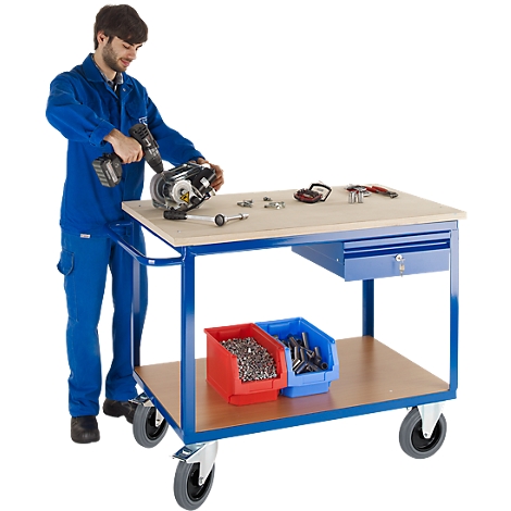 A man in blue overalls working on a workbench with tools. The table is on wheels.