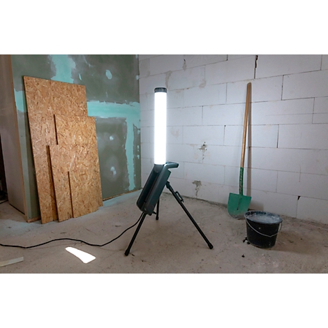 Une lampe de chantier lumineuse sur un trépied dans une pièce. A côté, pelle, seau et planches de bois.