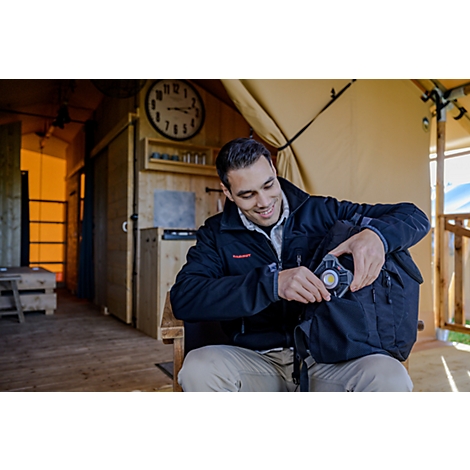 Un homme souriant porte une veste noire et est assis sur une chaise en bois. Il examine un sac à dos noir tout en fixant un objet rond et jaune dessus. En arrière-plan, on voit une cabane en bois avec une grande horloge et quelques appareils élect...