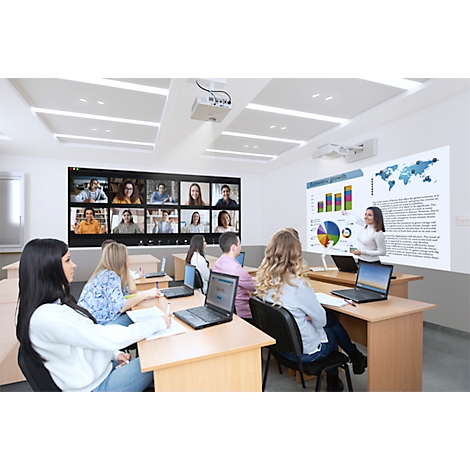 Dans une salle de classe, une femme se tient devant un écran affichant des schémas et une carte du monde. Il y a une vidéoconférence sur un écran séparé.