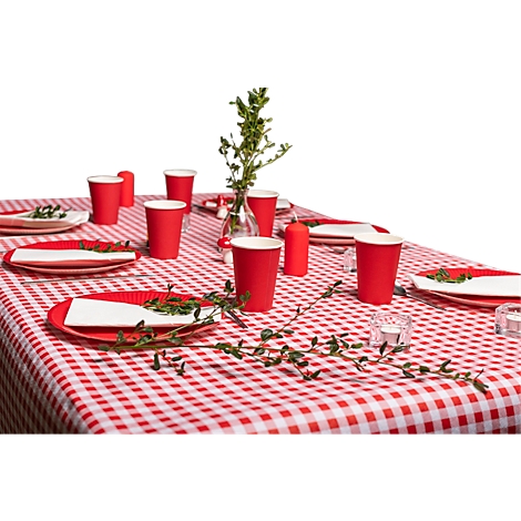 Une table est dressée avec une nappe à carreaux rouges. Des assiettes, des gobelets, des bougies et des branches vertes sont sur la table.