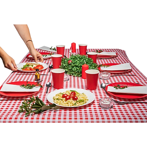 Une table dressée avec une nappe à carreaux rouges et blancs, des assiettes de nourriture, des gobelets rouges et des bougies, une main posant une assiette.