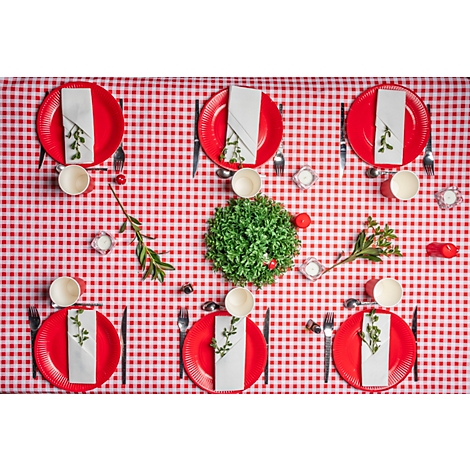 Table avec nappe à carreaux rouges. Assiettes rouges avec serviettes, couverts et tasses. Décoré de verdure et de bougies.