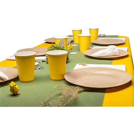 Ein festlich gedeckter Tisch mit gelben Bechern, braunen Tellern und Tischdecke mit Grün und Gelb. Ein Küken und Zweige dekorieren den Tisch.
