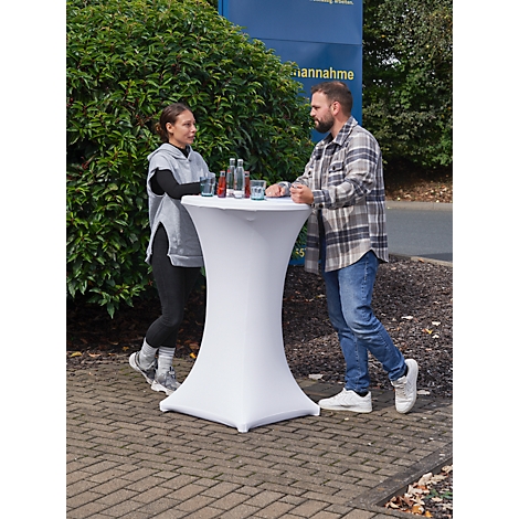 Two people stand at a white high table with drinks. In the background a building with writing.