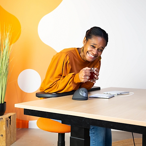 A smiling woman drinks tea at a desk. She is wearing an orange top and sitting at a standing desk.