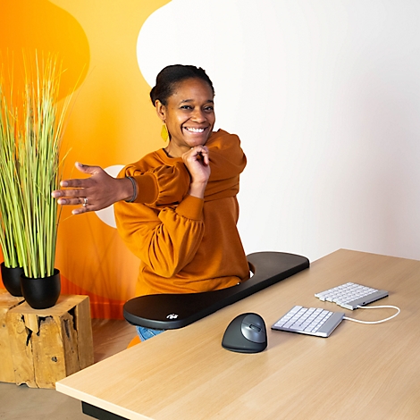 Smiling woman stretching at a desk, ergonomic accessories in front: mouse, keyboard, tray.