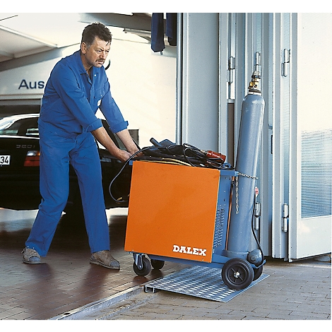 Un homme en salopette bleue pousse un chariot de soudage avec un coffre orange et une bouteille de gaz.