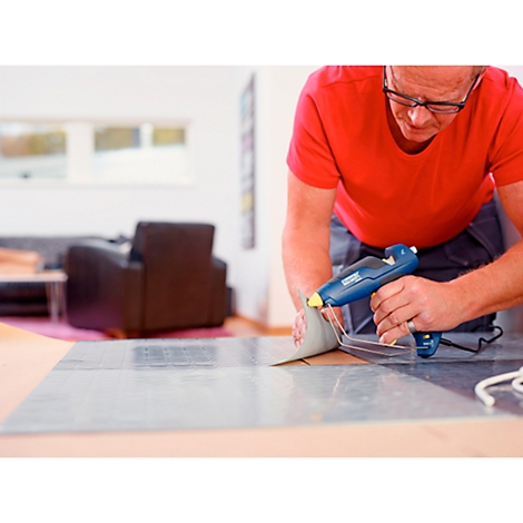A man glues something onto a plate with a hot glue gun. He is wearing a red t-shirt and glasses.