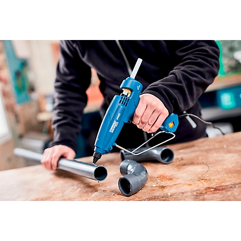 Person using a hot glue gun to connect a pipe section with a corner. The blue gun is in focus.