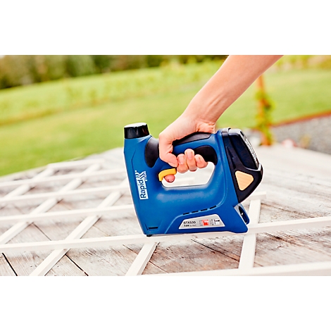 Person using a blue stapler to attach wooden strips, with a green meadow in the background.